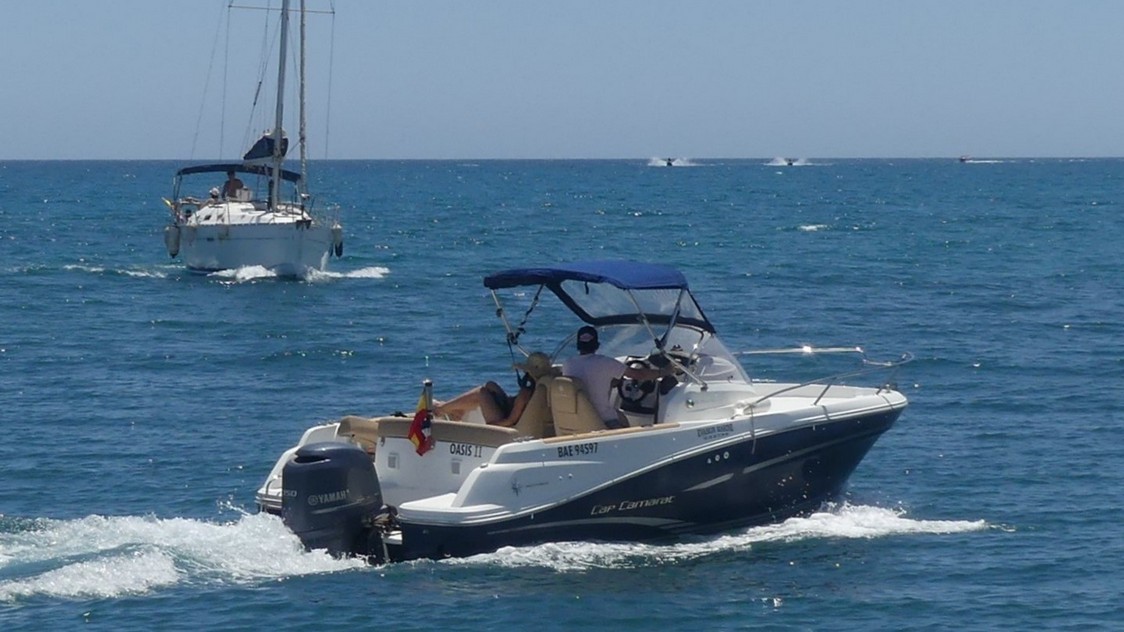 Lancha de motor con velero blanco que se acerca y al fondo motos acuáticas